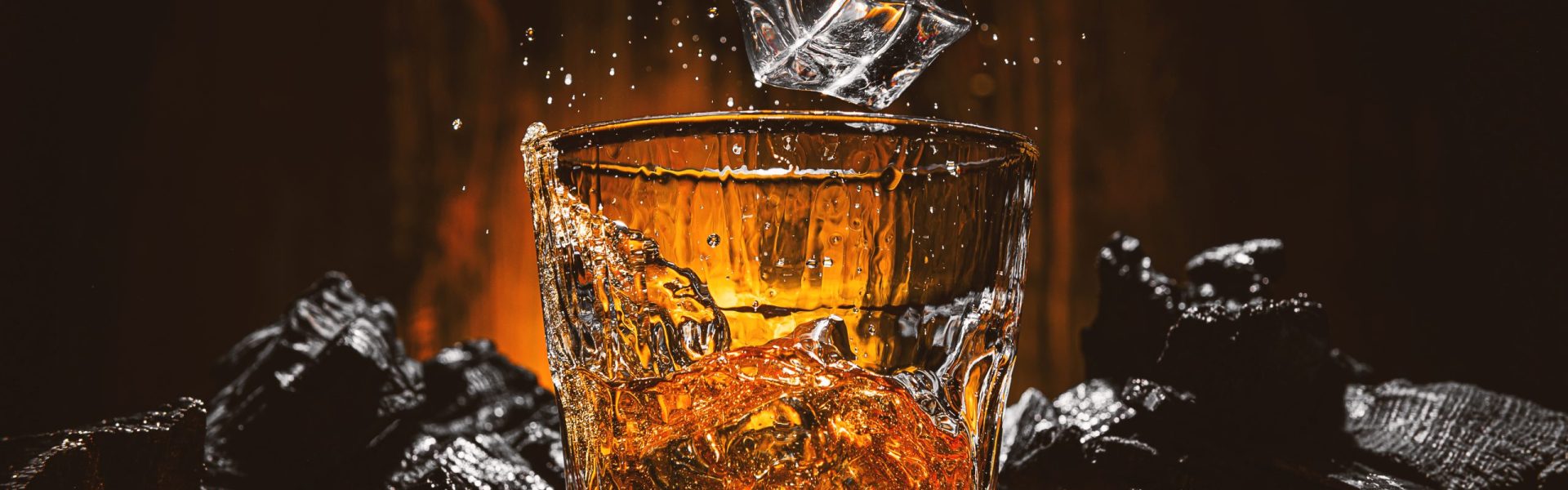 ice cubes fall into a glass with a brown alcoholic drink. splashes from falling ice fly in different directions